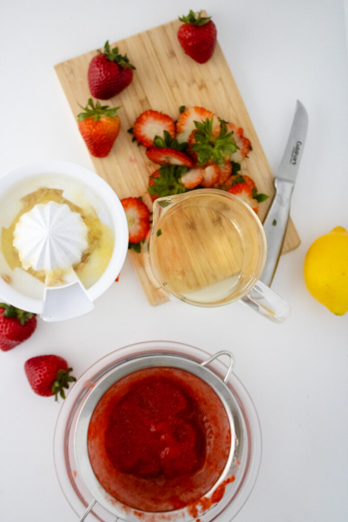 strawberry puree, lemon juice, and an empty pitcher sitting on top of a wooden chopping board with sliced strawberries and a knife