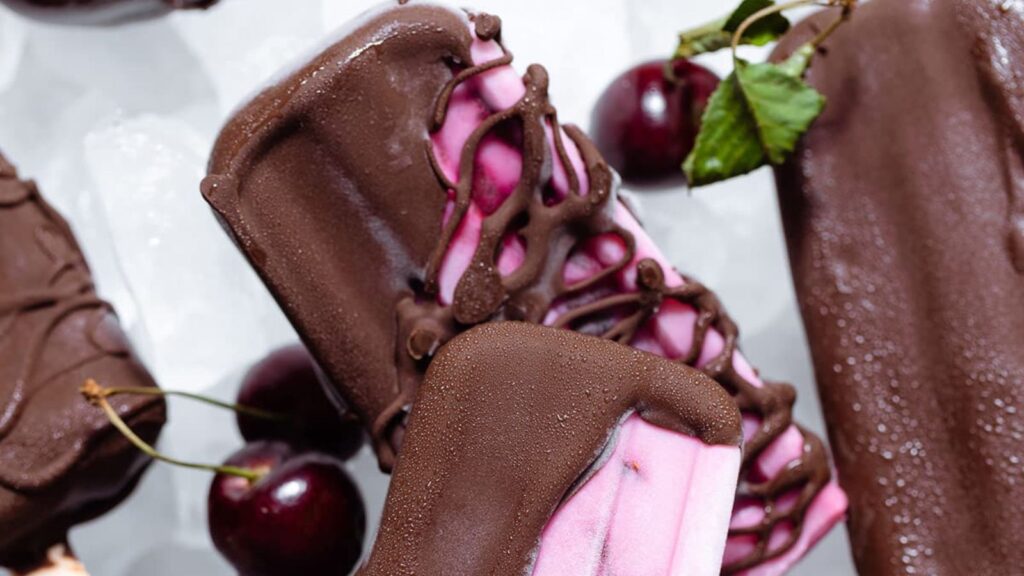 21 Delightful Homemade Popsicle Recipes 8 Pink cherry popsicles partially covered with dark chocolate on a white serving plate with ice.