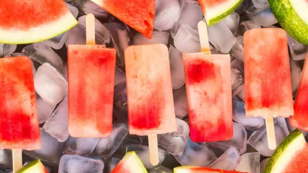 21 Delightful Homemade Popsicle Recipes 13 Watermelon popsicles and slices of fresh watermelon on a tray covered in ice from overhead.