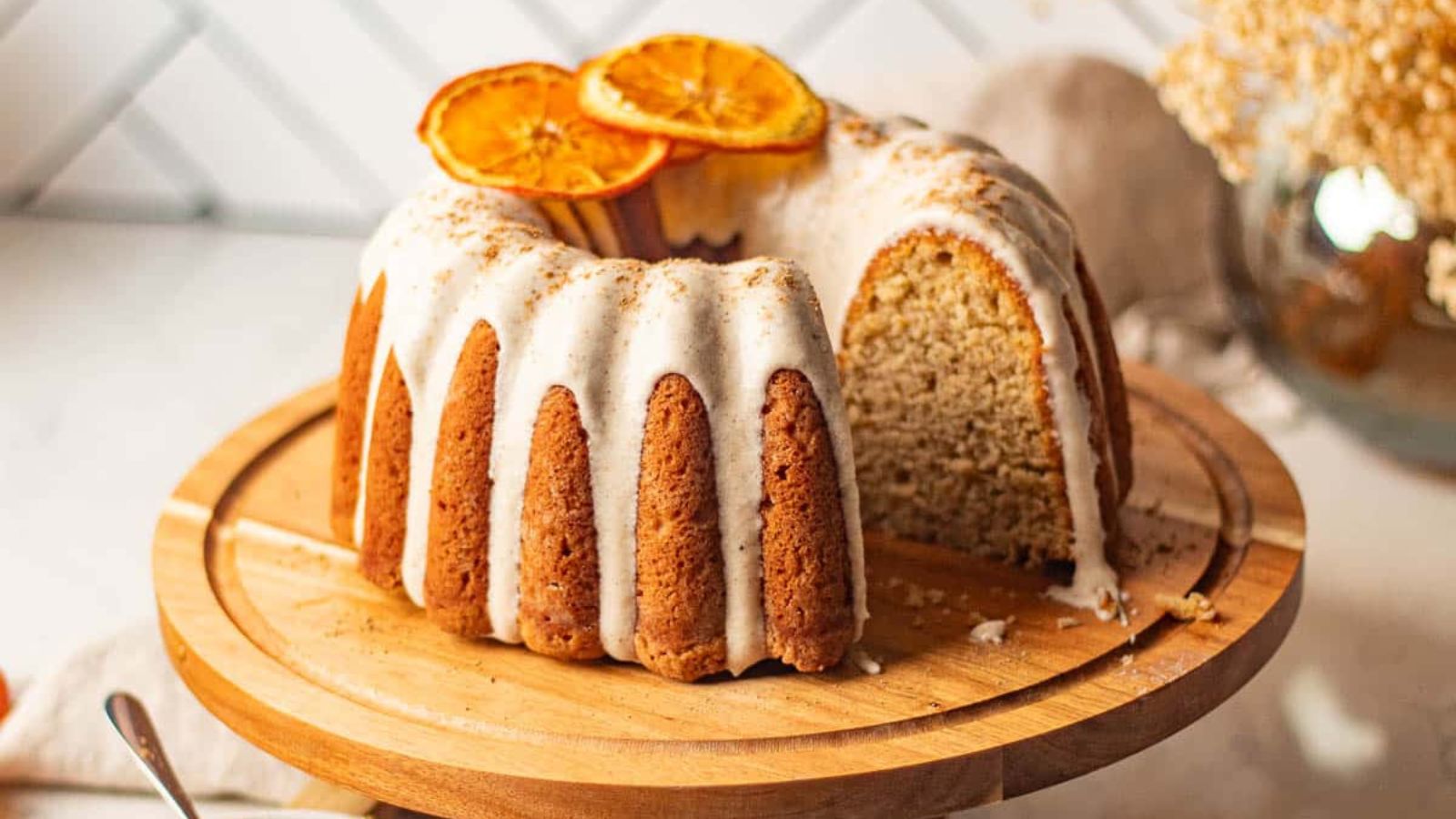A chai bundt spice cake with a slice cut out, revealing its moist, spiced interior, topped with cream cheese frosting and dried orange slices, presented on a wooden cake stand.