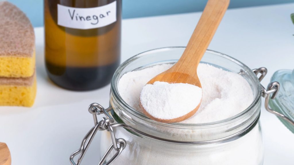 A jar filled with baking soda and a wooden spoon rests on a countertop alongside a bottle of vinegar and cleaning sponges. DIY cleaning method using natural products