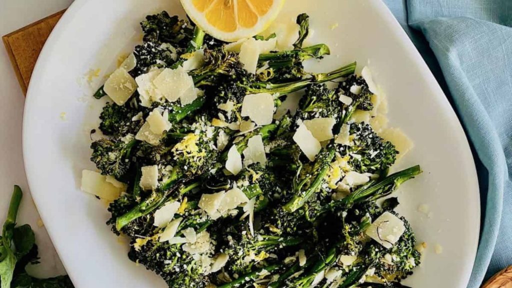 Overhead of white platter filled with broccoli topped with parmesan and lemon juice and serving utensils and blue napkin to the side.