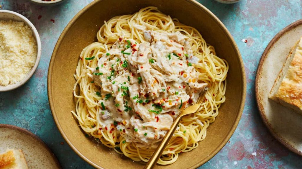 Shredded chicken in a white wine cream sauce over a bed of angel hair pasta in a bowl on a turquoise background.