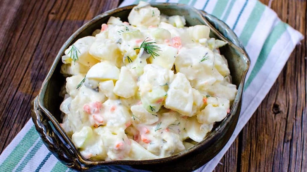 24 Potato Recipes You’ll Never Get Tired Of 19 Dill Pickle Potato Egg Salad in a dark green bowl on a blue, green and white striped fabric napkin on a wooden table.